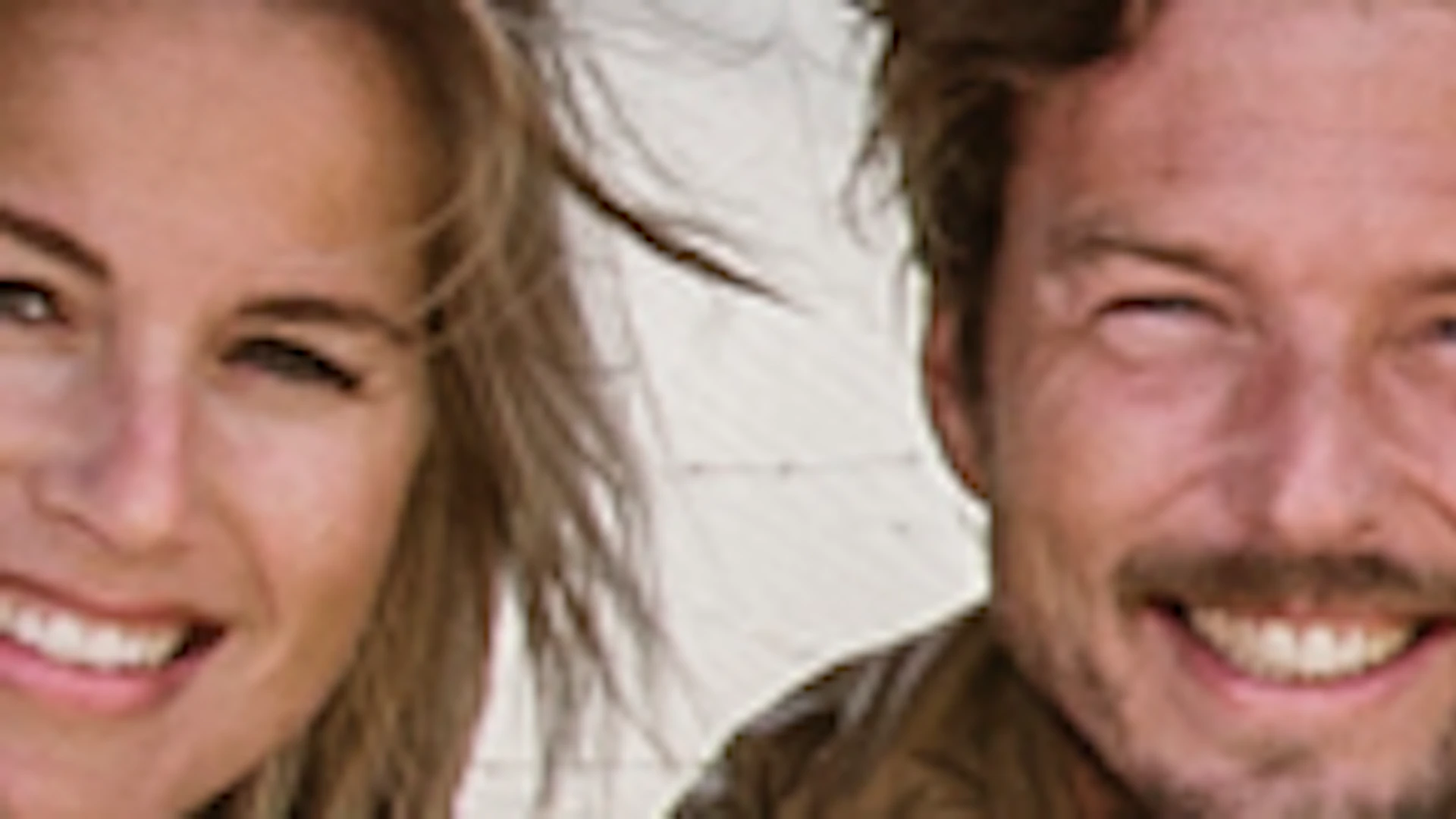 Two people sit in a small, camouflaged electric vehicle with upward opening door, parked on a brick road near sandy dunes.