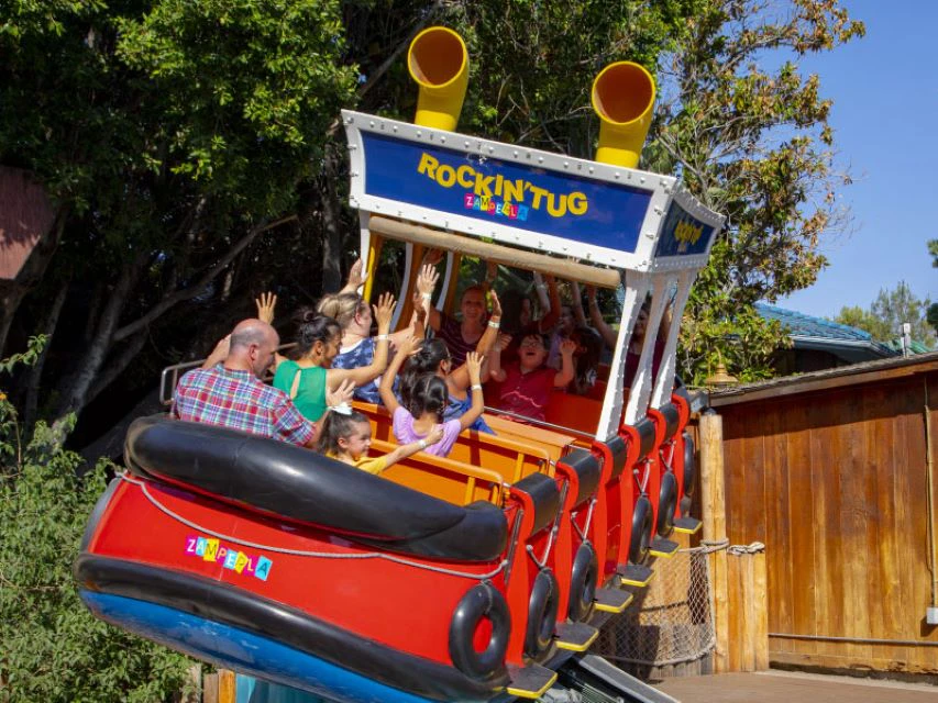Children and adults with raised hands enjoying a ride on the Rockin' Tug amusement park attraction. Trees in the background.