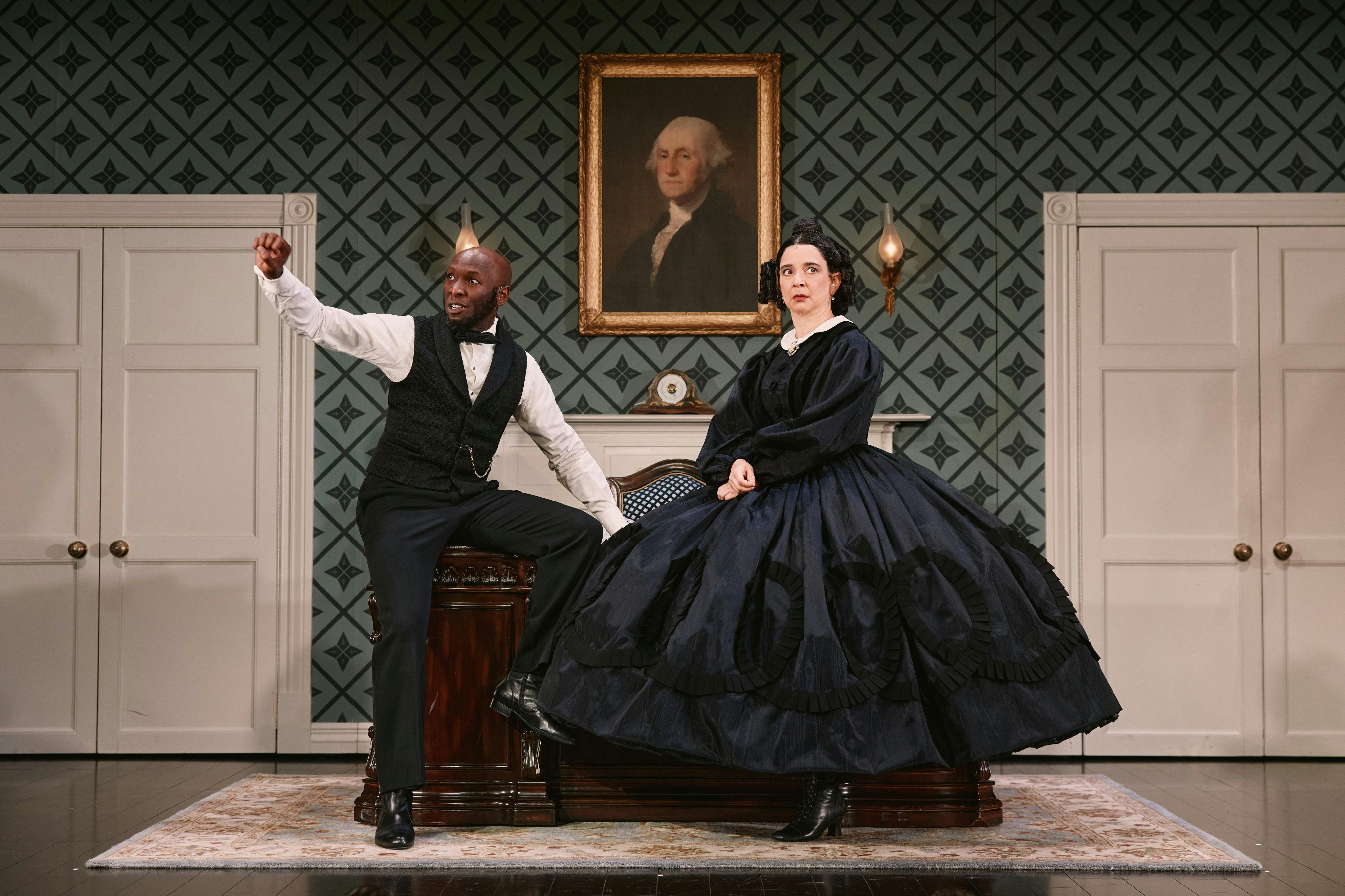 Two actors in period costumes performing on a stage with a portrait of George Washington on the wall behind them.