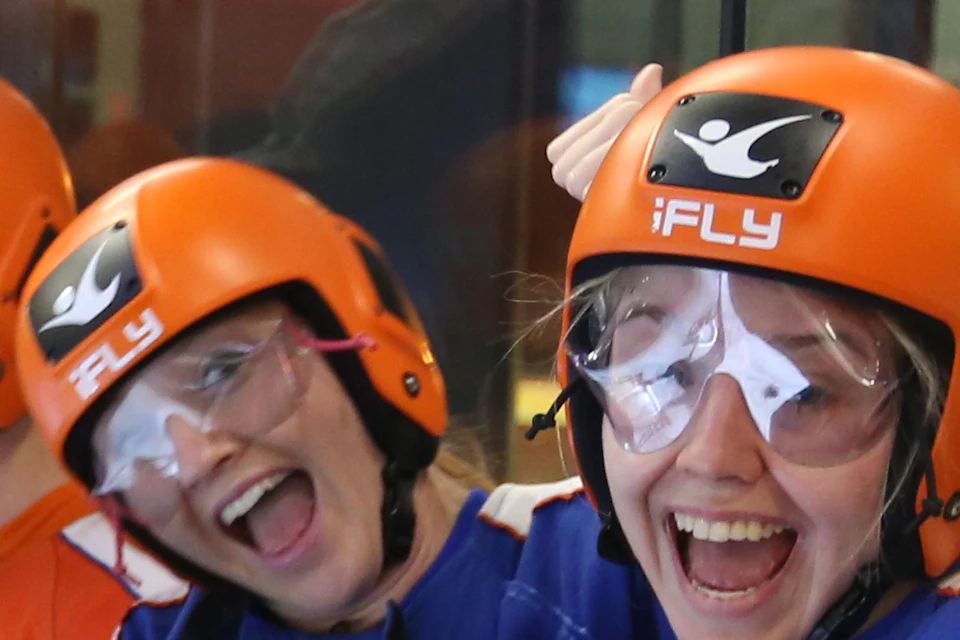Group of four people in helmets and jumpsuits sitting and smiling in an indoor skydiving center, giving thumbs up.
