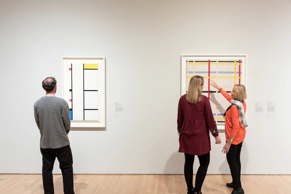 Two people observe abstract art in a gallery, with framed geometric paintings on a white wall.