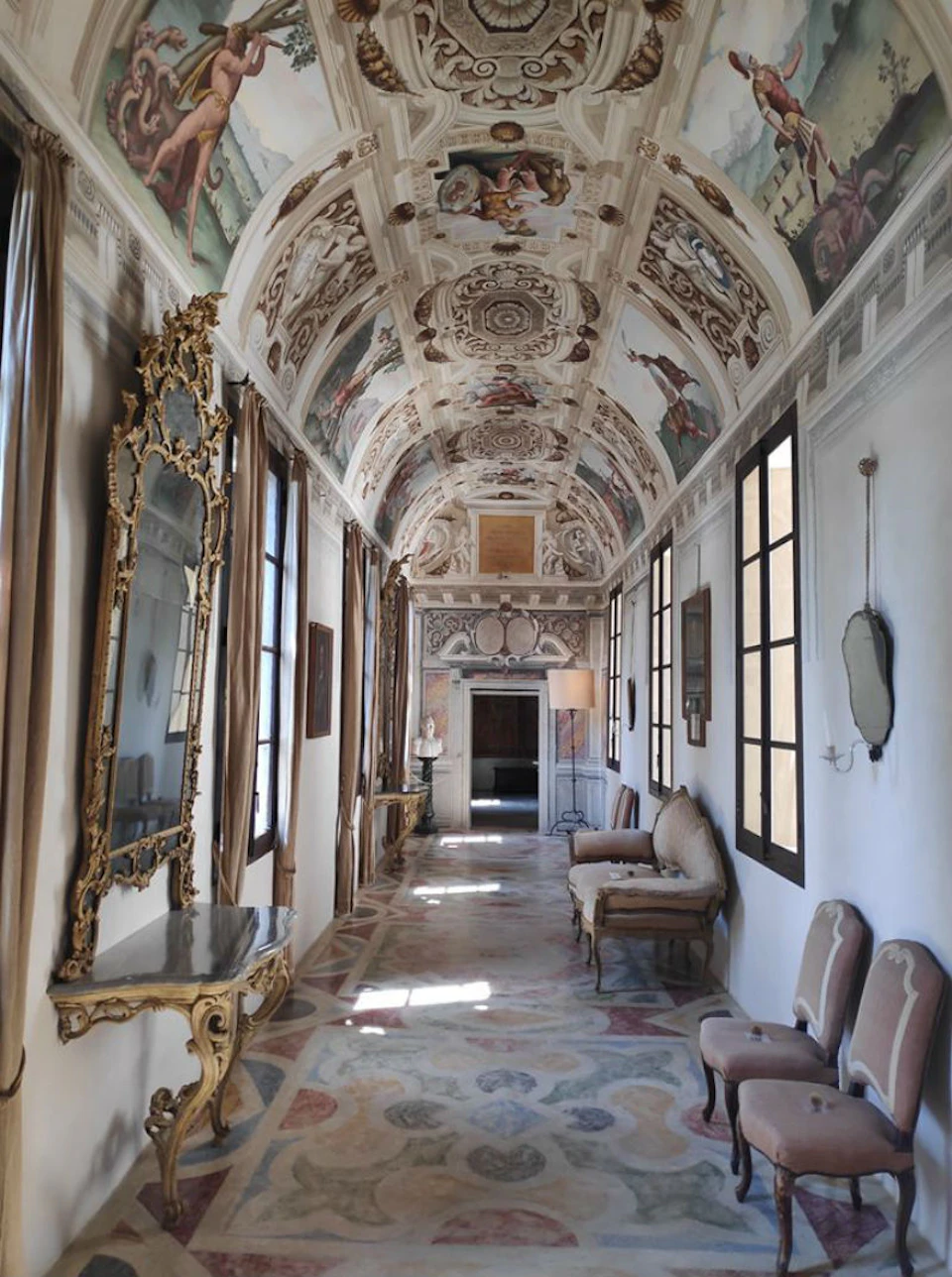 Ornate hallway with intricate ceiling frescoes, gilded mirrors, antique furniture, and large windows with natural light streaming in.