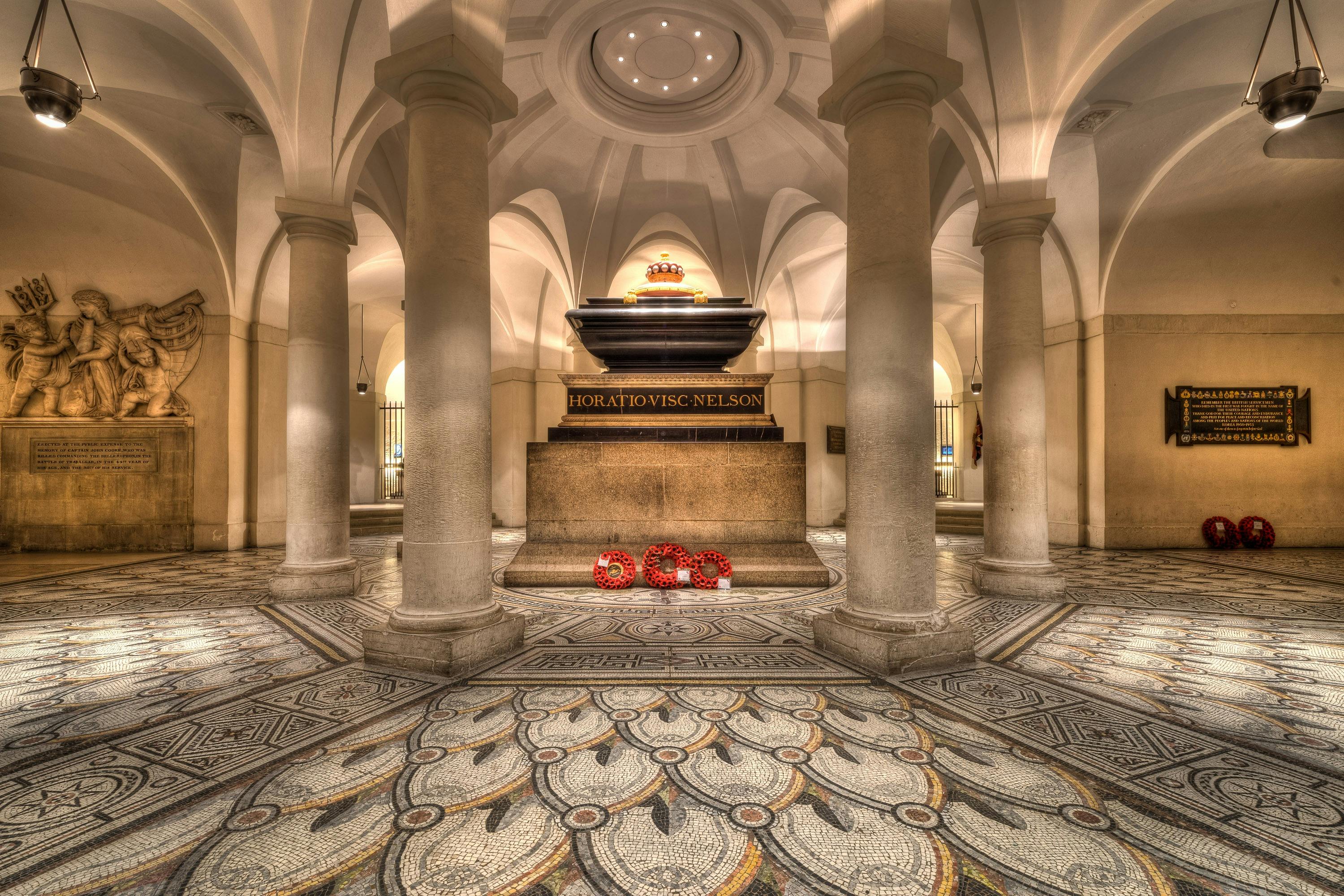 A grand hall with pillars, ornate flooring, and a central stone sarcophagus surrounded by wreaths and marked "Horatio Viscount Nelson."
