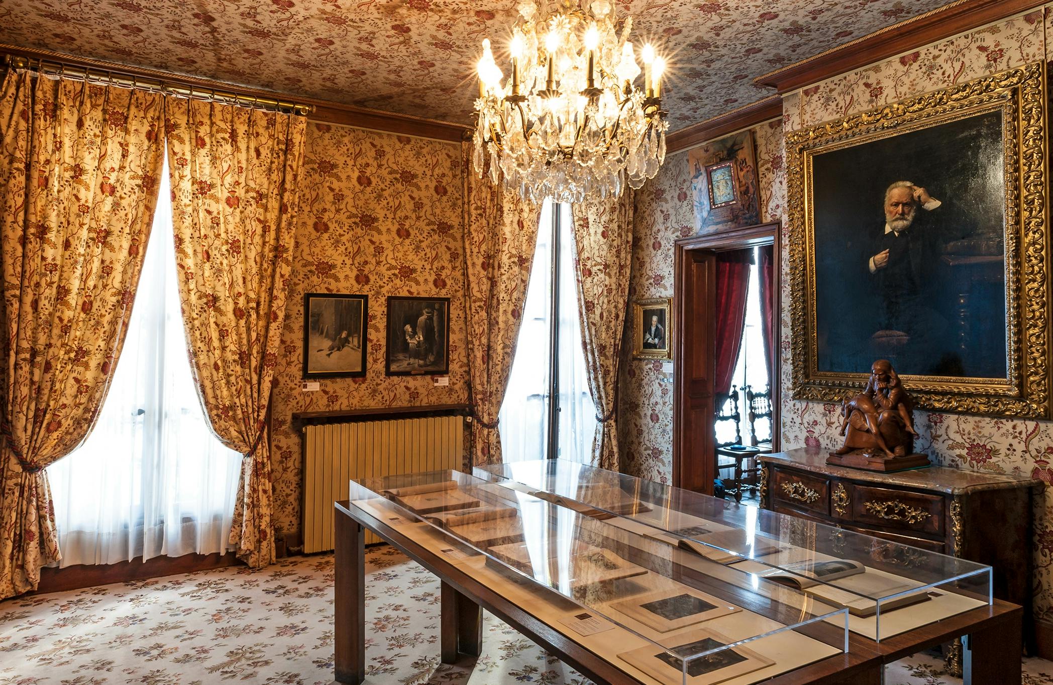Habitació adornada amb paper pintat de flors, candelabro, retrats emmarcats, mobles antics i una taula d'exposició amb documents.