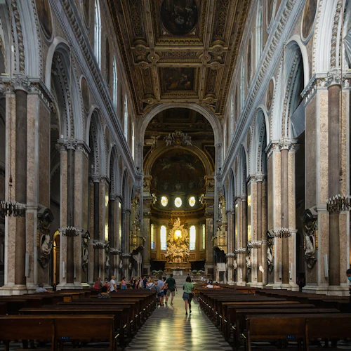 Insidan av en stor katedral med höga pelare, utsmyckade bågar, träbänkar och människor som går mot ett omsorgsfullt dekorerat altare.