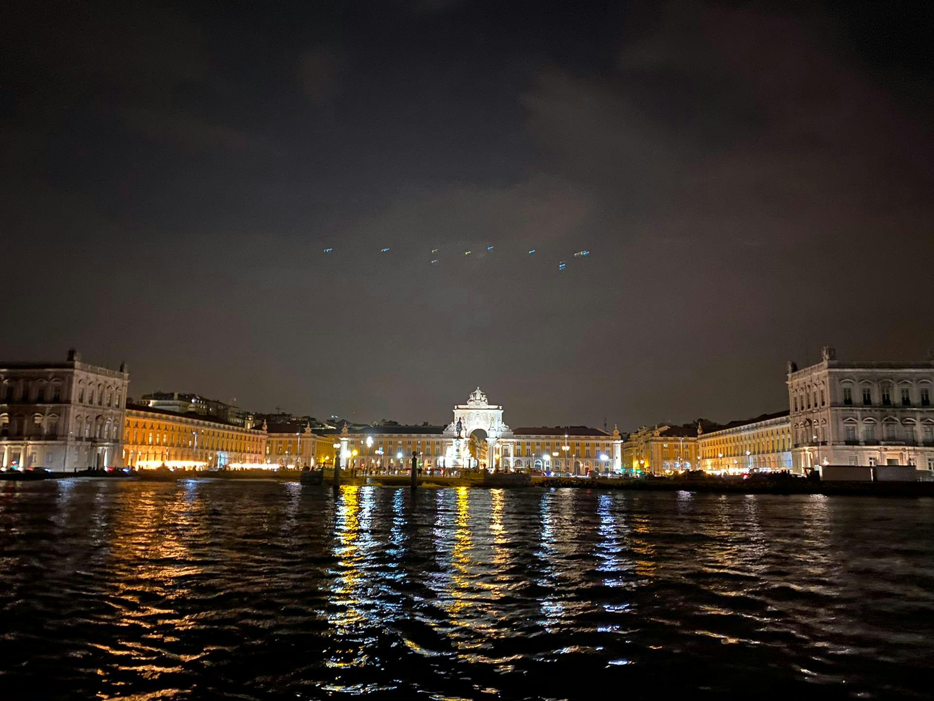 夜景是一个灯火通明的海滨城市广场，中心有一个华丽的拱门，映照在水面上。