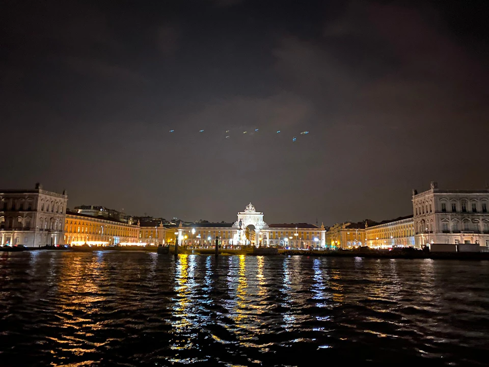 Nighttime view of a lit-up waterfront city square with an ornate archway in the center, reflected in the water's surface.
