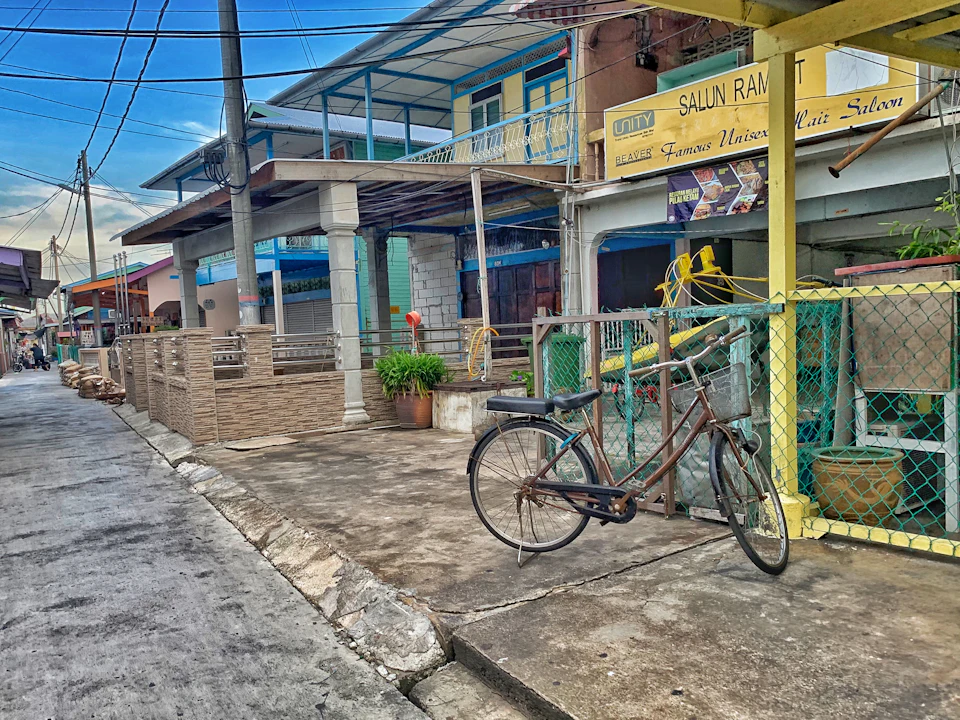 A bicycle is parked on a sidewalk in front of a rustic building with a "Salun Ram" sign,​ next to a fenced area.