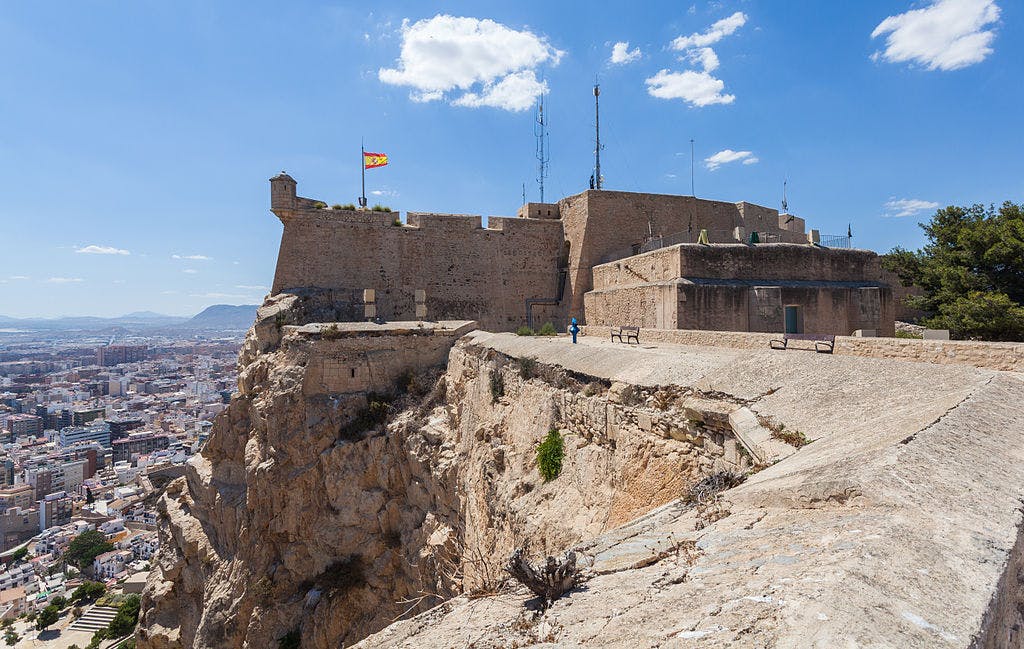 Stone fortress with a Spanish flag on a cliff, overlooking a cityscape under a clear blue sky. A person walks near the fortress.