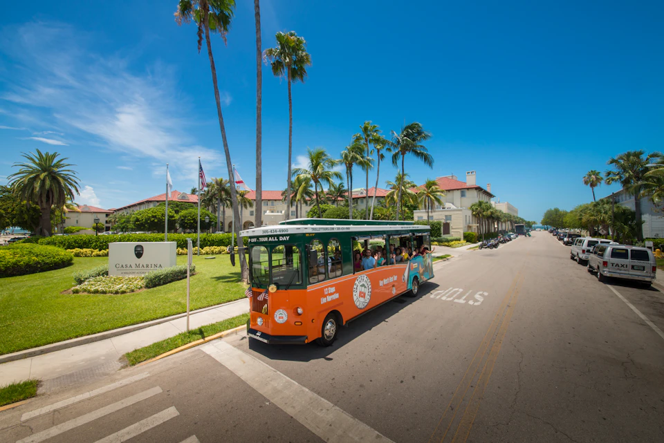 An orange and green tour trolley with passengers on a palm-lined street in front of "Casa Marina" under a clear blue sky.