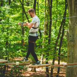 Person som går på en trädtoppsbana i en skog, bär säkerhetssele och håller i en guidekabel.