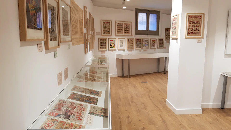 A room with wood flooring displaying framed artwork and documents on the walls and under glass cases.