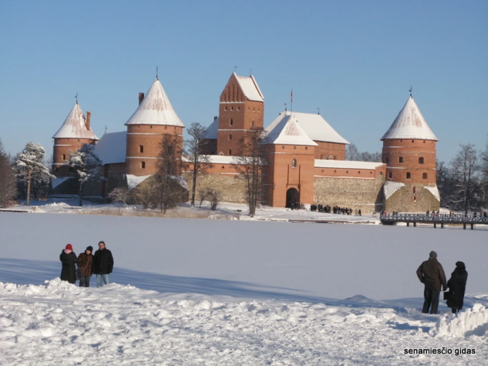 People standing in snow near a large brick castle with several round towers, all topped with snow, under a clear blue sky.
