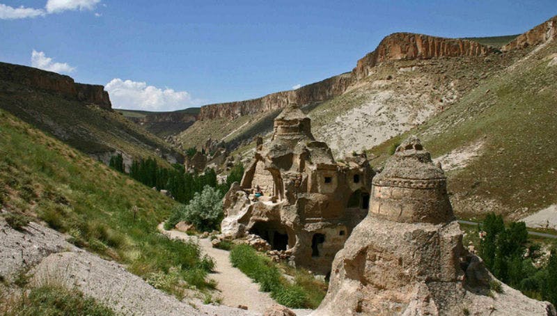 Rocky landscape featuring cave dwellings with arched openings, surrounded by greenery, steep cliffs, and a clear blue sky.