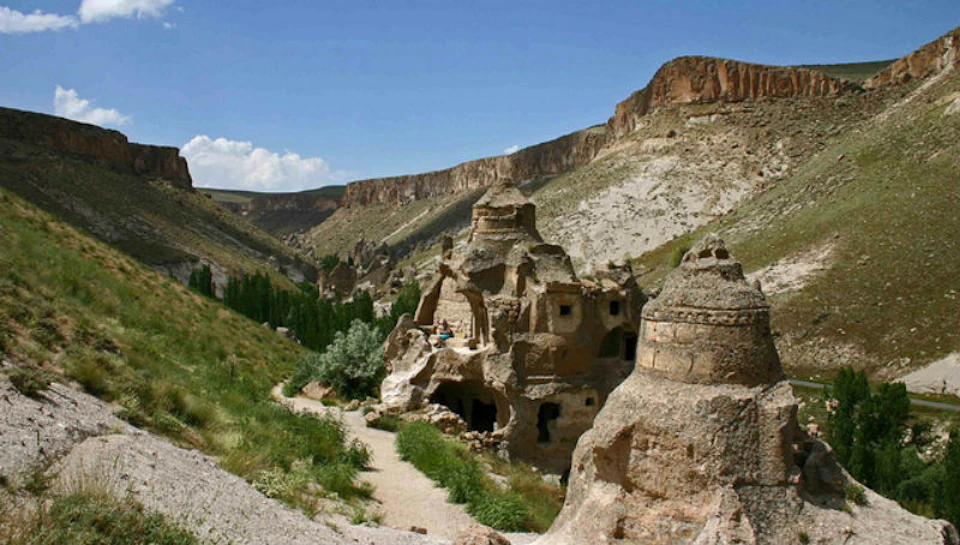 Rocky landscape featuring cave dwellings with arched openings, surrounded by greenery, steep cliffs, and a clear blue sky.