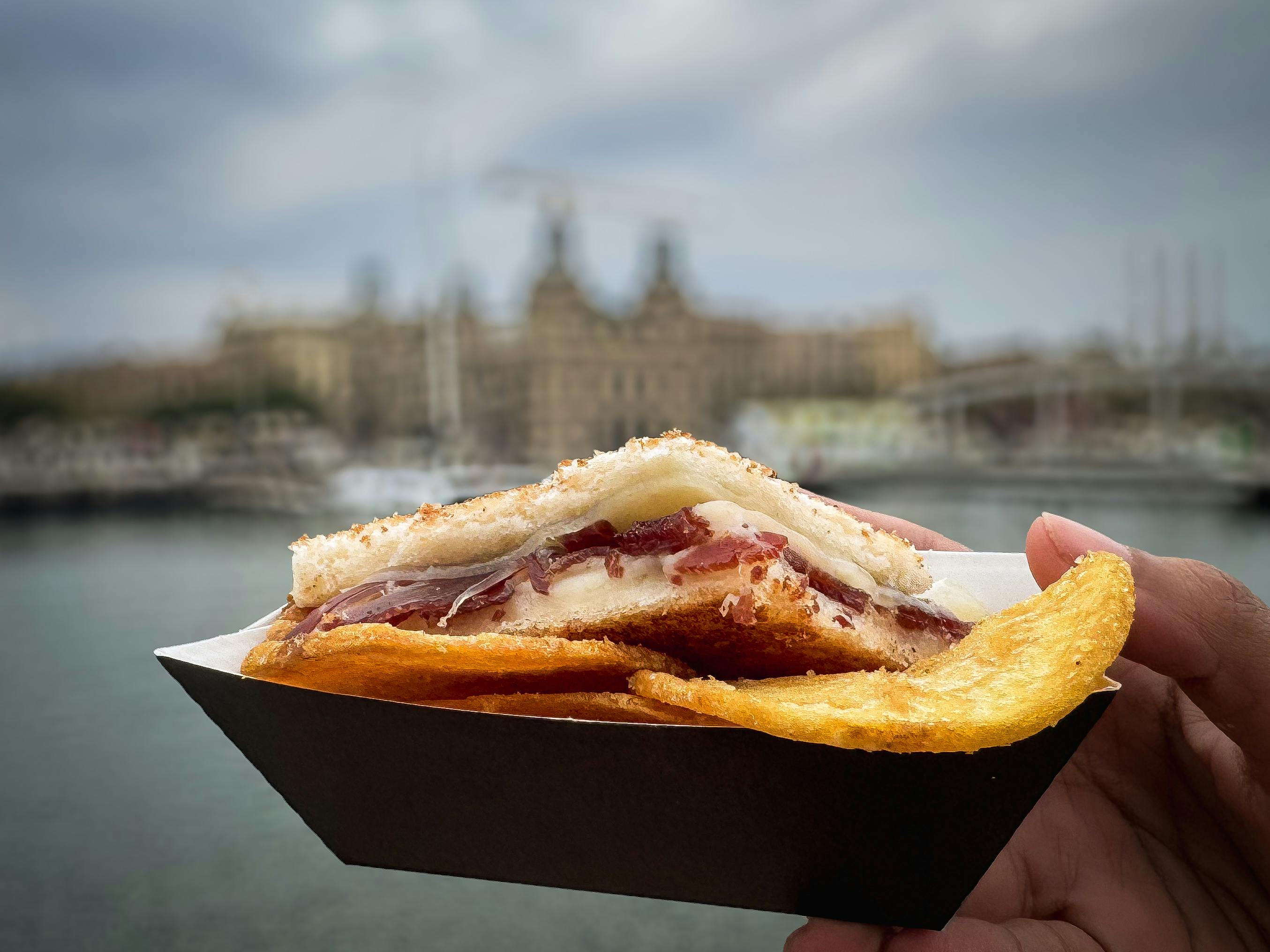 Ein Sandwich mit Schinken und Käse, serviert mit Kartoffelchips in einem Papiertablett, das im Freien in der Nähe einer Uferpromenade und von Gebäuden steht.