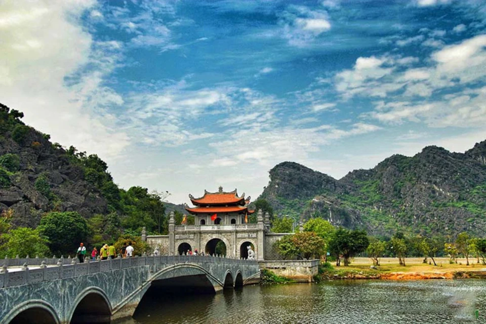 Bridge over water leading to a traditional Asian gate with a red roof, surrounded by trees and rocky hills under a partly cloudy sky.
