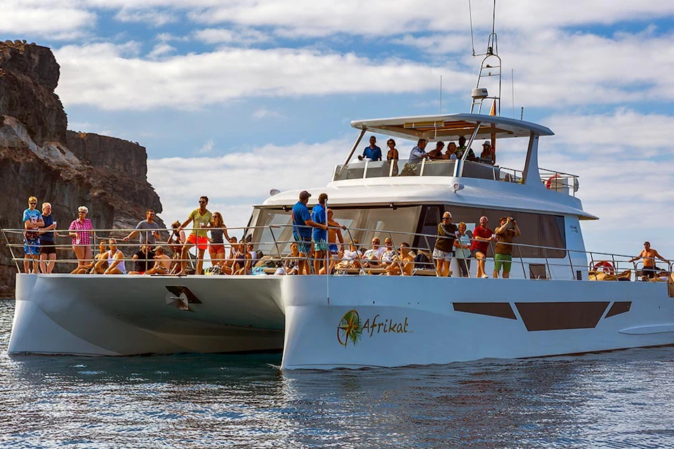 A large catamaran with people on board enjoying the sunny day on the water, with rocky cliffs and a partly cloudy sky in the background.