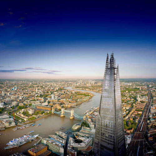 Flygfoto över The Shard skyskrapa i London med Themsen, Tower Bridge och stadsbilden under en klarblå himmel.