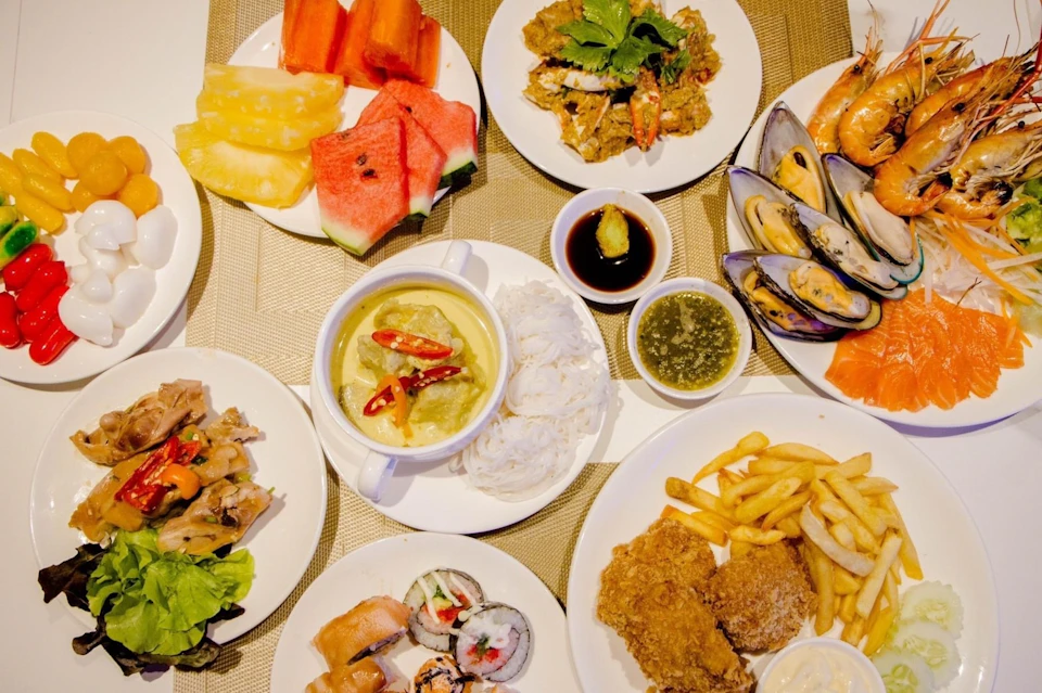 A variety of dishes including watermelon, pineapple, mussels, sushi, fried chicken, fries, noodle soup, and dipping sauces.