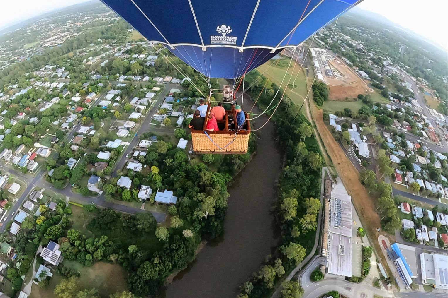 Ein Heißluftballon mit Passagieren, die über ein Wohngebiet, einen Fluss und die umliegenden Grünflächen fliegen.
