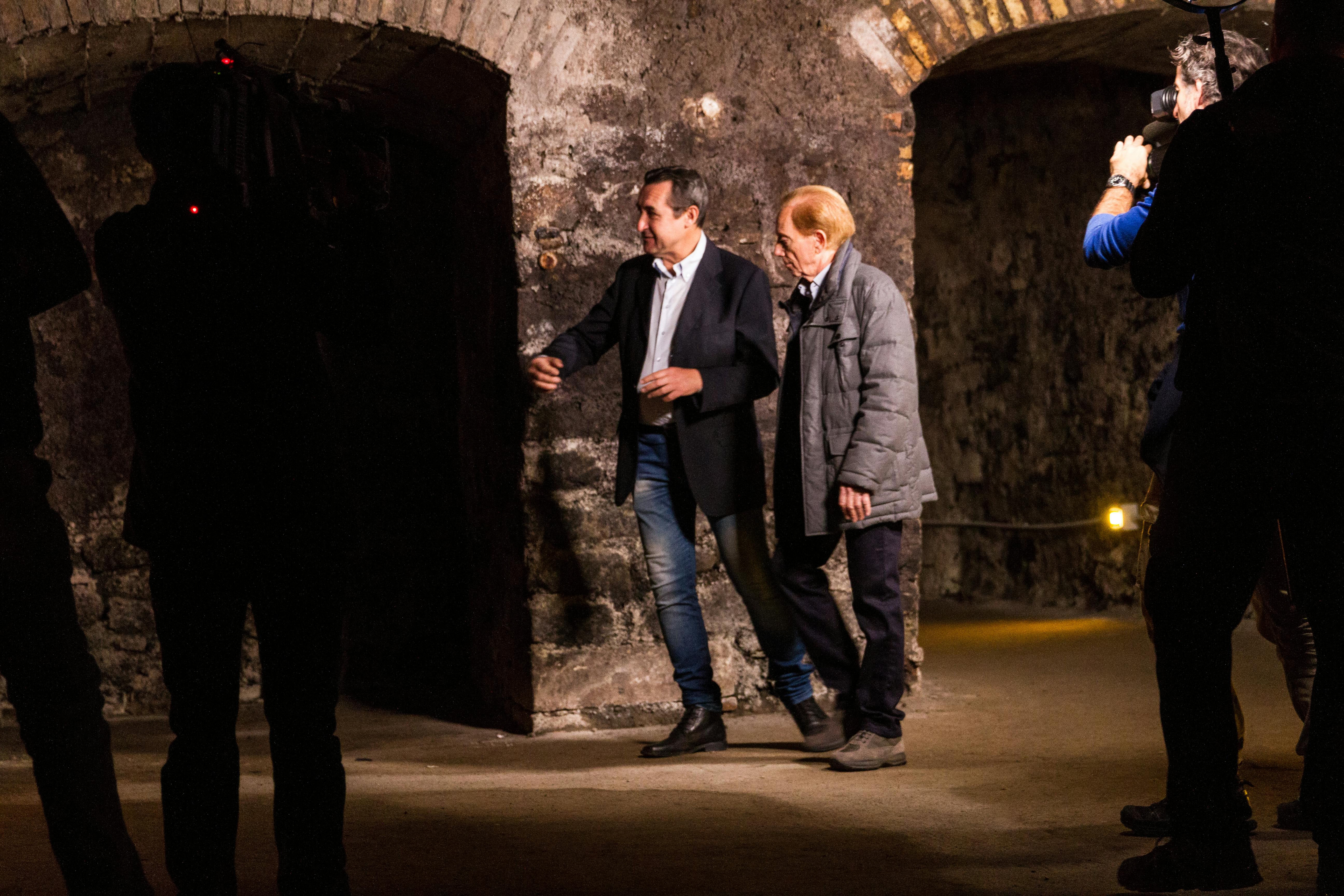 To mænd, der går i en svagt oplyst stengang med buede lofter; en mand i en blazer og jeans, den anden i en grå frakke.