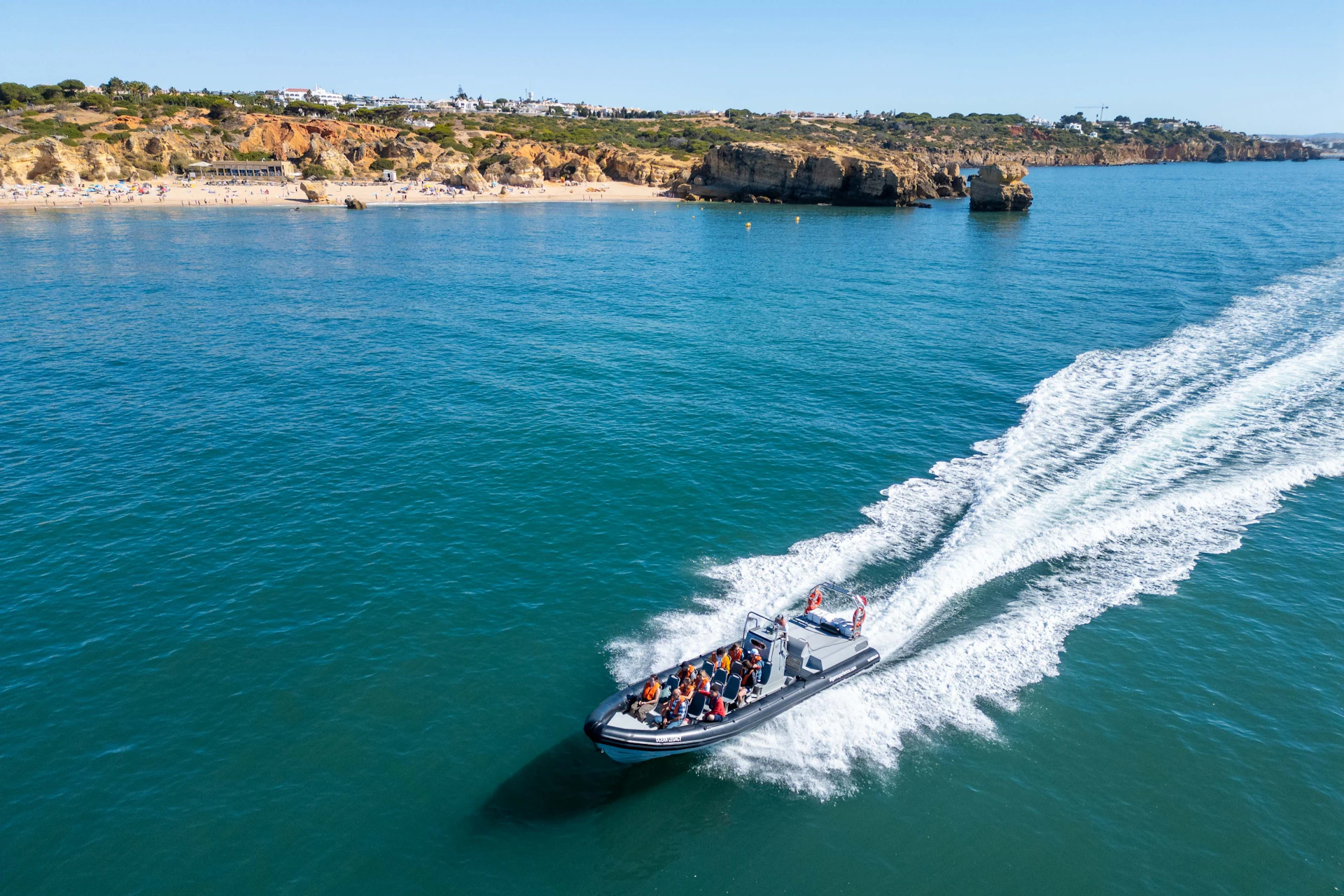 A speedboat with passengers cruising on blue water near a rocky coastline under a clear sky.