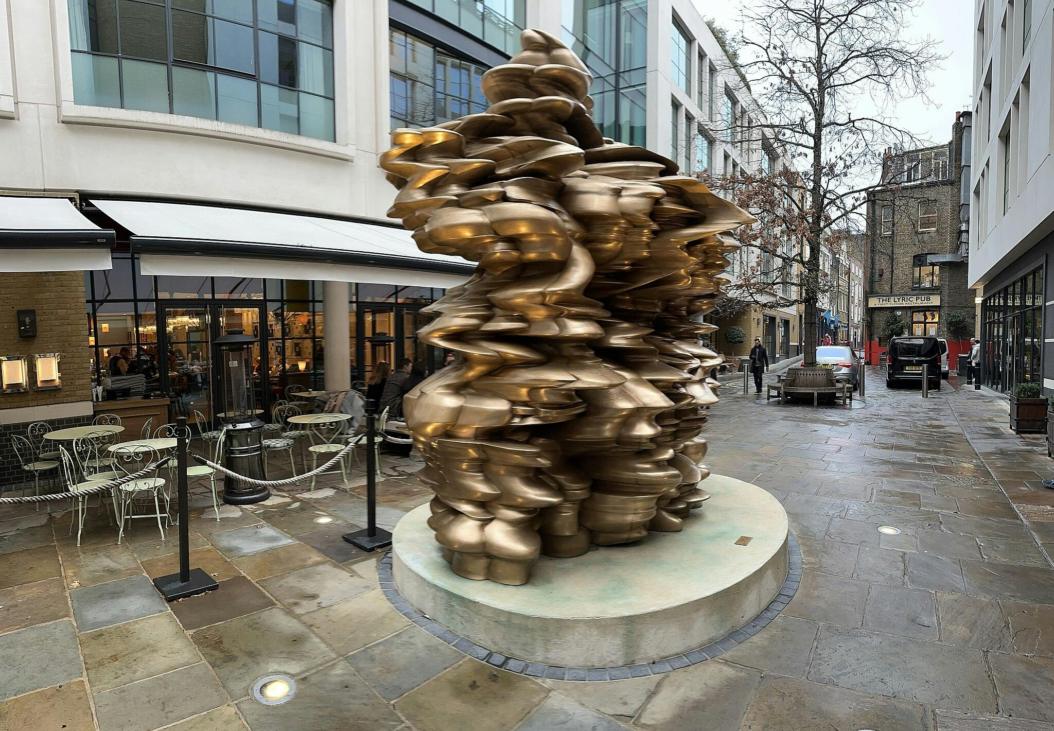 Bronzová abstraktní socha stojí ve venkovním městském prostředí s kavárnami, lavičkami a chodci. Budovy a obchody jsou v blízkosti.