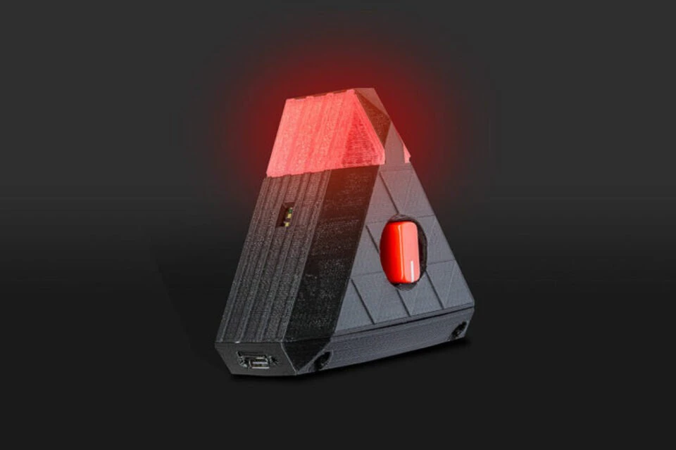 A black, pyramid-shaped electronic device with a glowing red top and a red button, set against a dark background.