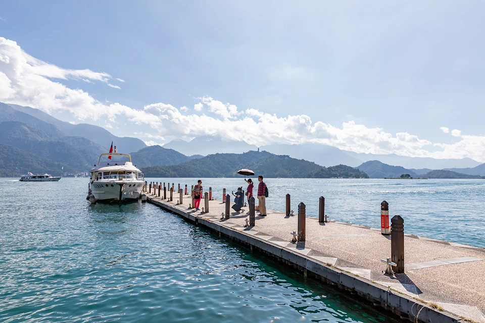 A dock extending into a lake with people walking, a moored boat, and mountains in the background under a sunny sky.