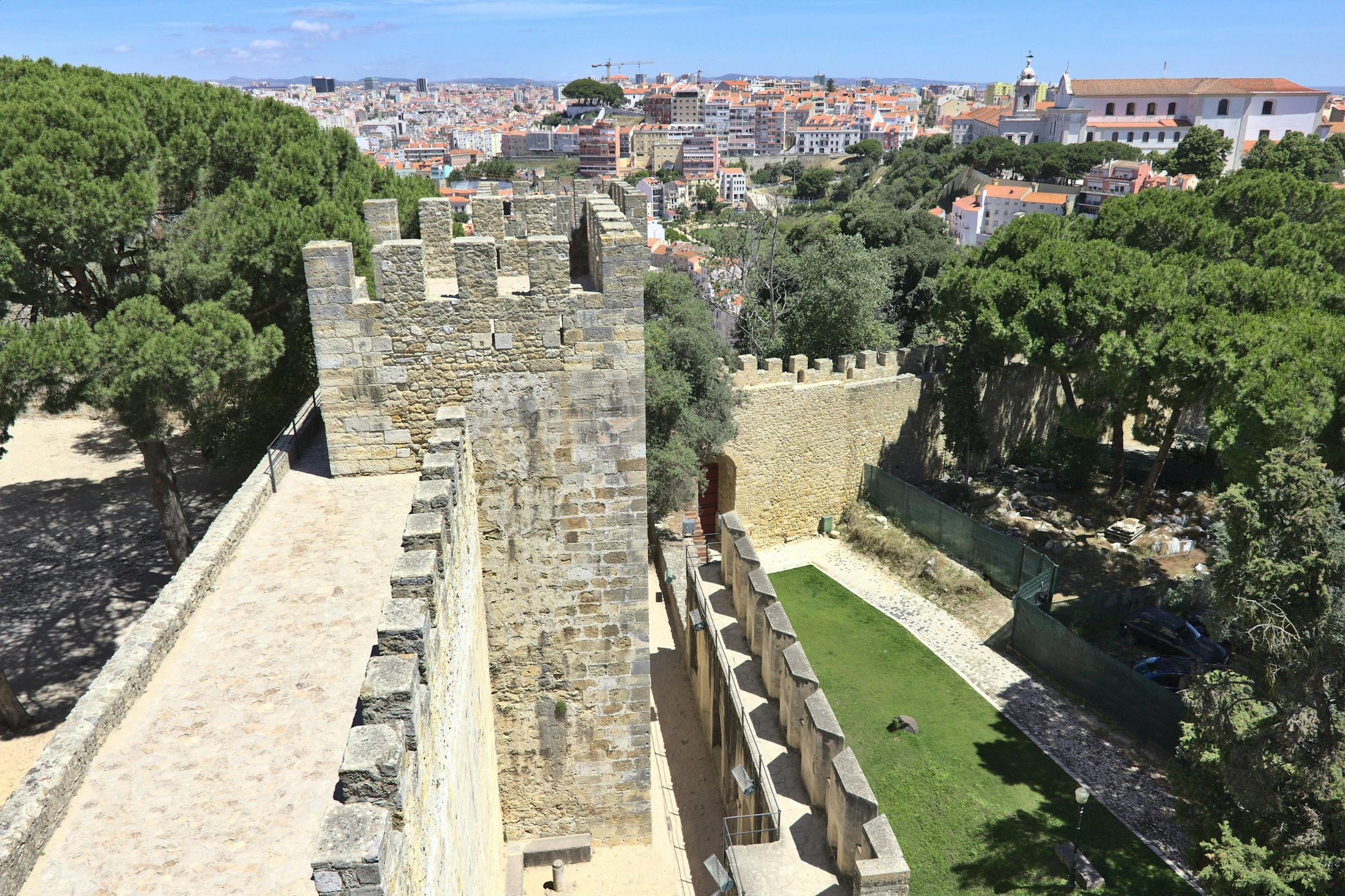 A historic castle wall with crenellations overlooks a cityscape and green trees under a sunny sky.