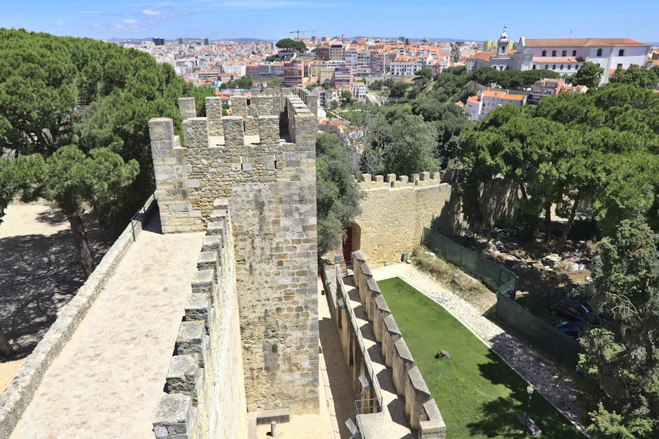 A historic castle wall with crenellations overlooks a cityscape and green trees under a sunny sky.
