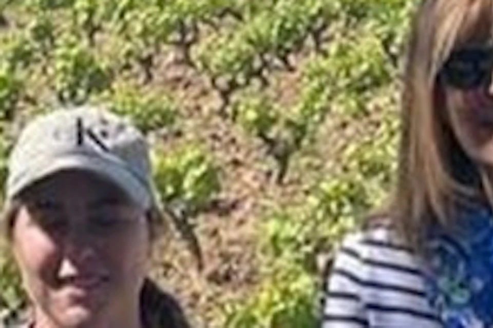 Four people stand smiling in front of a vineyard with rows of plants, surrounded by trees under a clear blue sky.