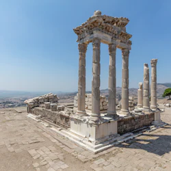 Ruiner av forntida stenkolonner och tempelstruktur med två personer som går i närheten under en klar himmel, med utsikt över ett avlägset landskap.