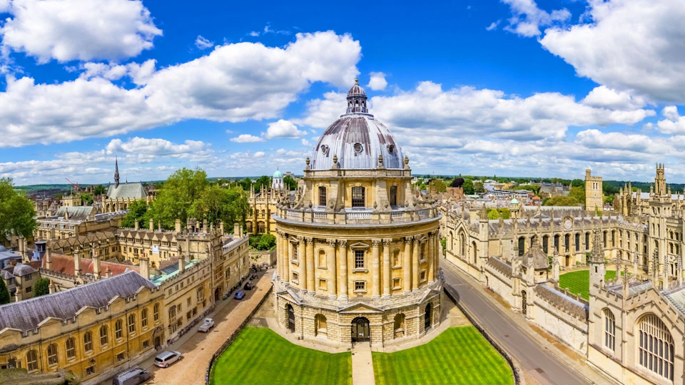 Aerial view of a historic domed building surrounded by green lawns and stone pathways, set under a blue sky with scattered clouds.