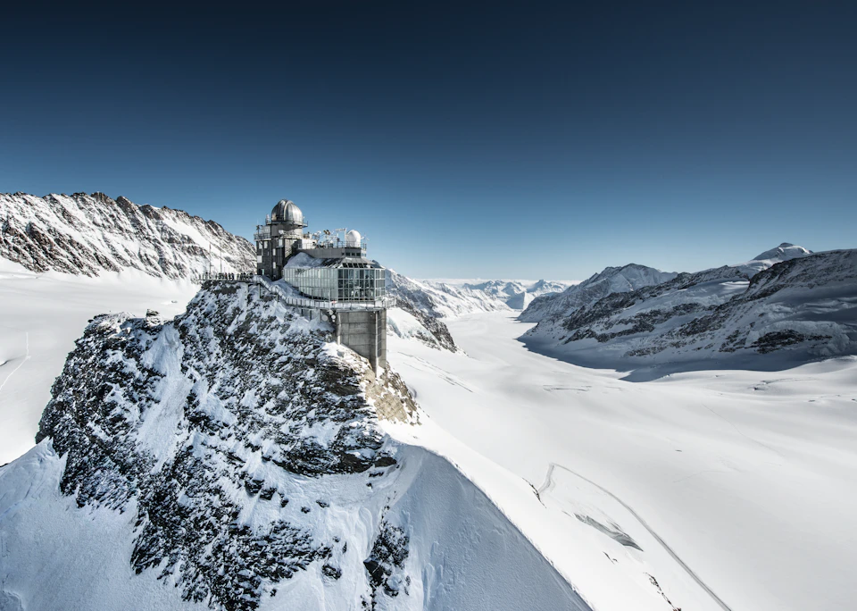 An observatory stands on a snowy mountain peak, overlooking a vast expanse of snow-covered valleys and distant mountain ranges.