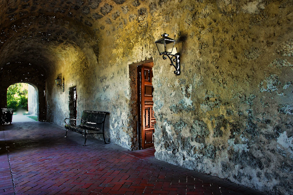 Stone corridor with arched ceiling, wrought iron bench, brick floor, wall lanterns, and a partially open wooden door.