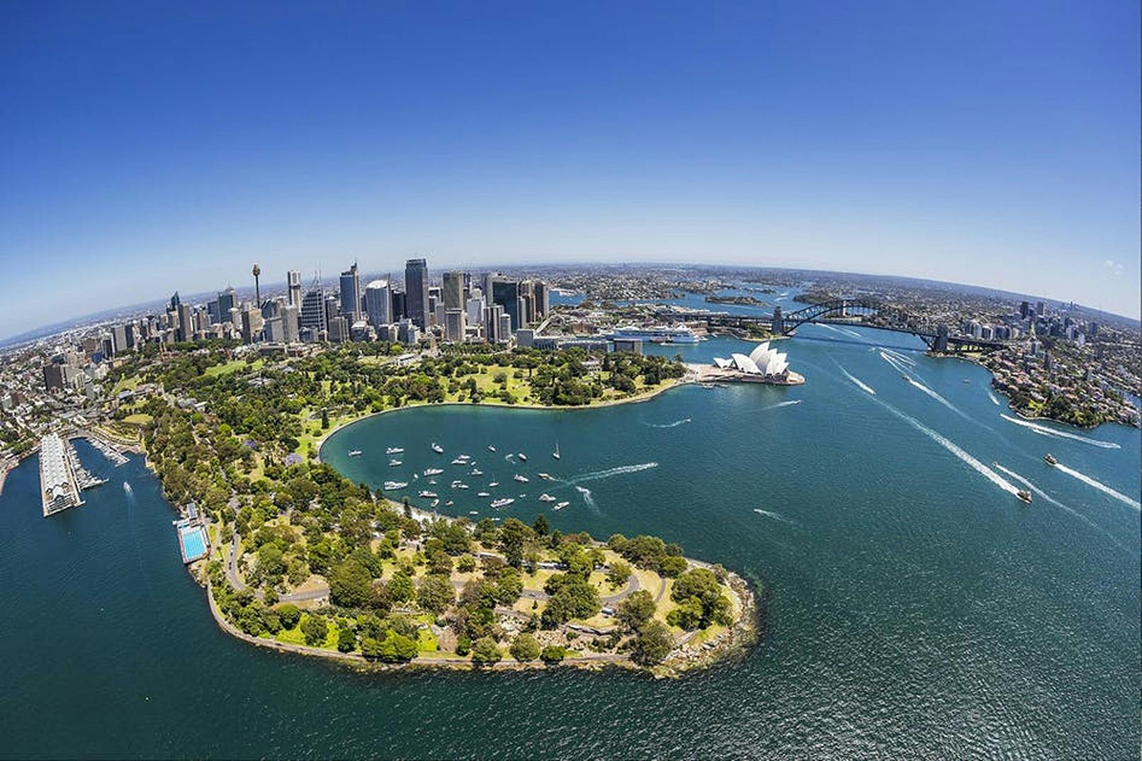 Aerial view of a city with a harbor, iconic buildings, green parkland, and numerous boats on the water under a clear blue sky.