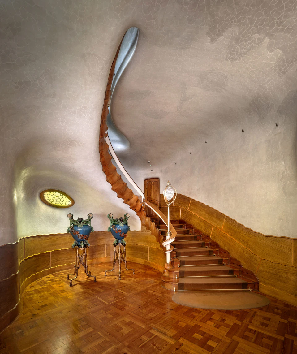 Curved wooden staircase with ornate railing in a uniquely designed room, featuring two decorative sculptures and an oval window.