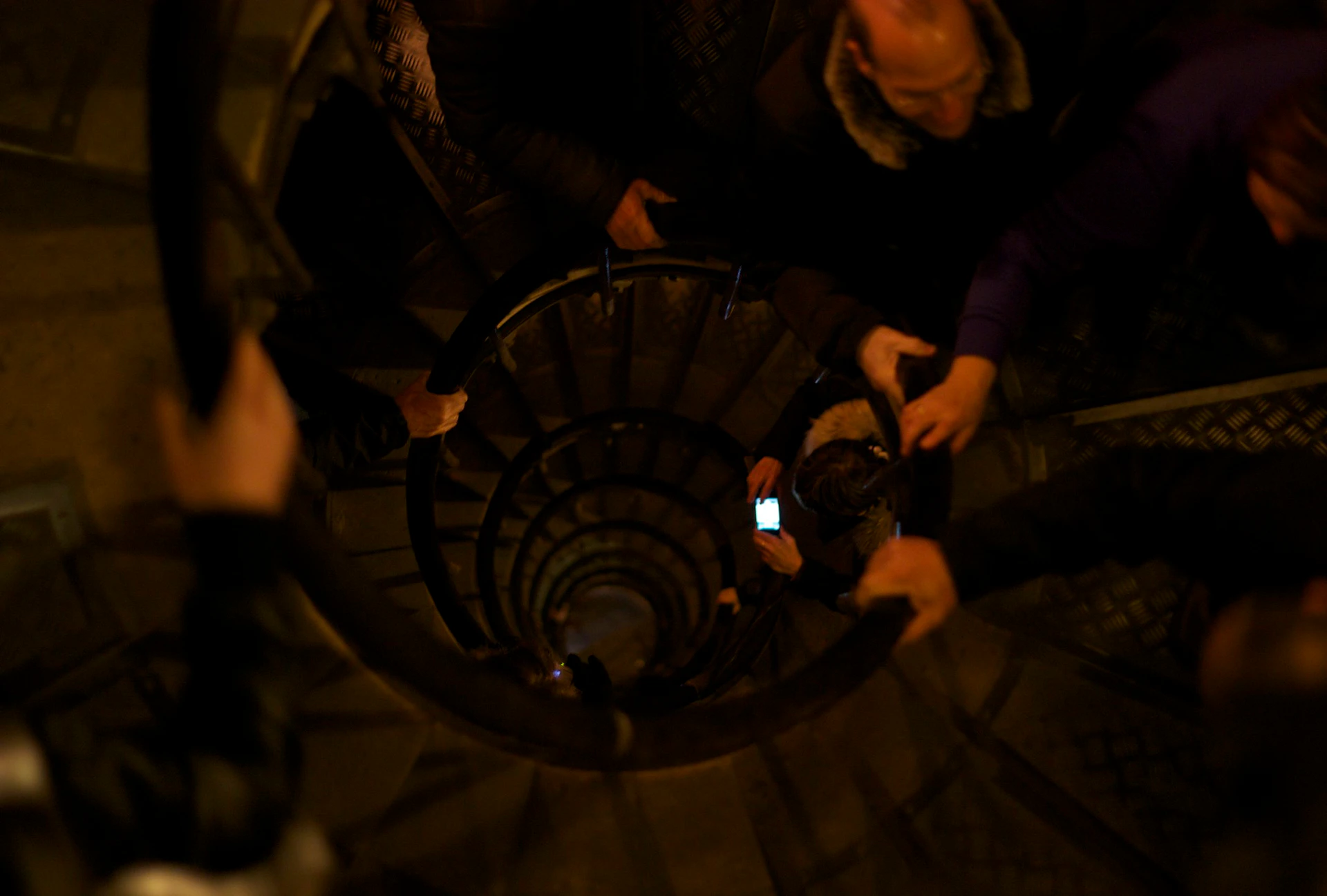 People descending a spiral staircase, with one person holding a lit smartphone in the center.