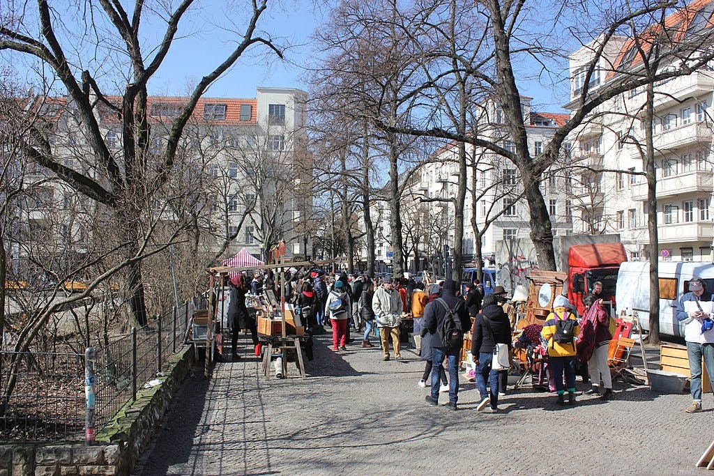 People walking and shopping at an outdoor market with stalls, trees, and residential buildings in the background on a sunny day.