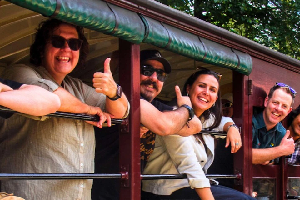 A group of people leaning out of the windows of a vintage train car, smiling and gesturing enthusiastically, with a conductor nearby.