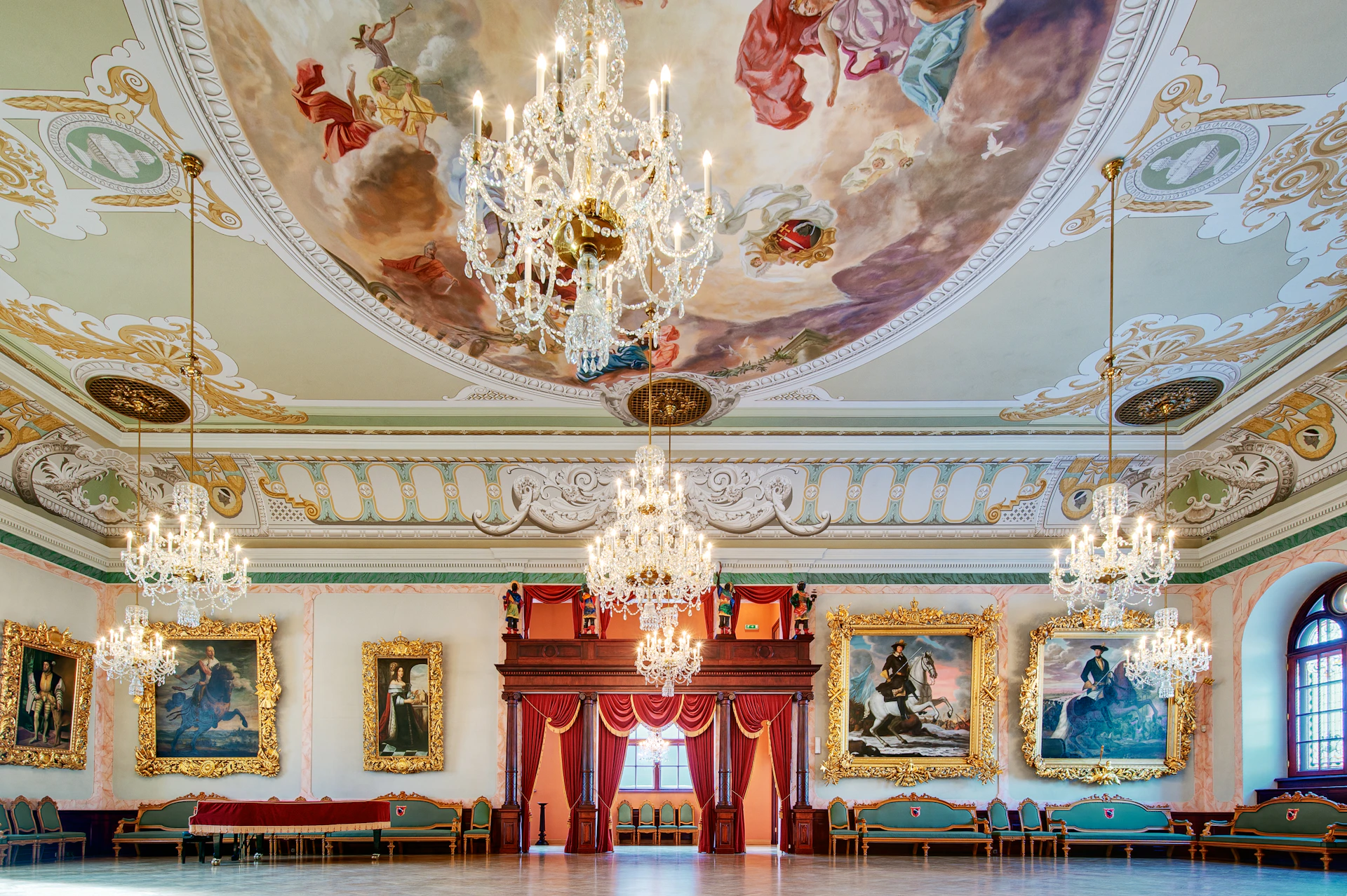 Ornate ballroom with chandeliers, large paintings, a red draped stage, and a ceiling mural depicting figures in the sky.