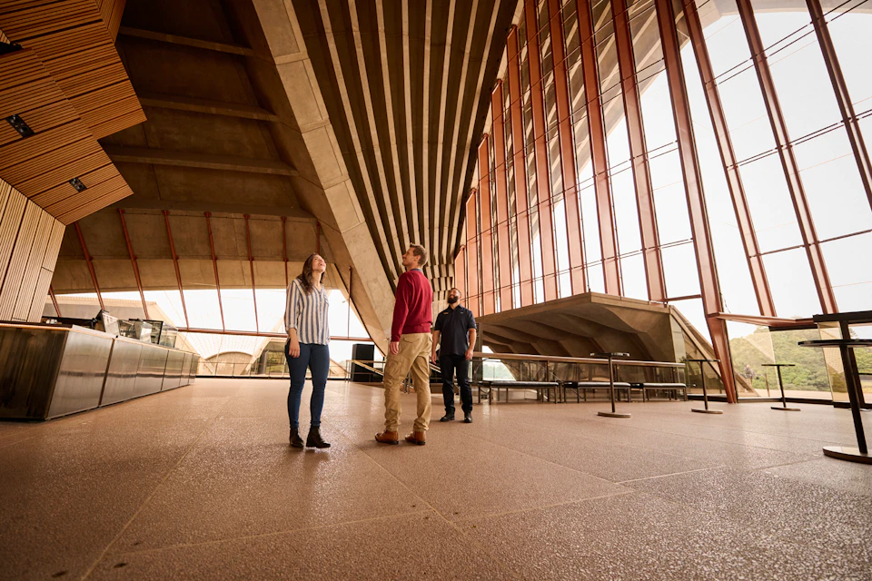 Three people stand in a spacious architectural interior with high ceilings, concrete beams, and large vertical windows.