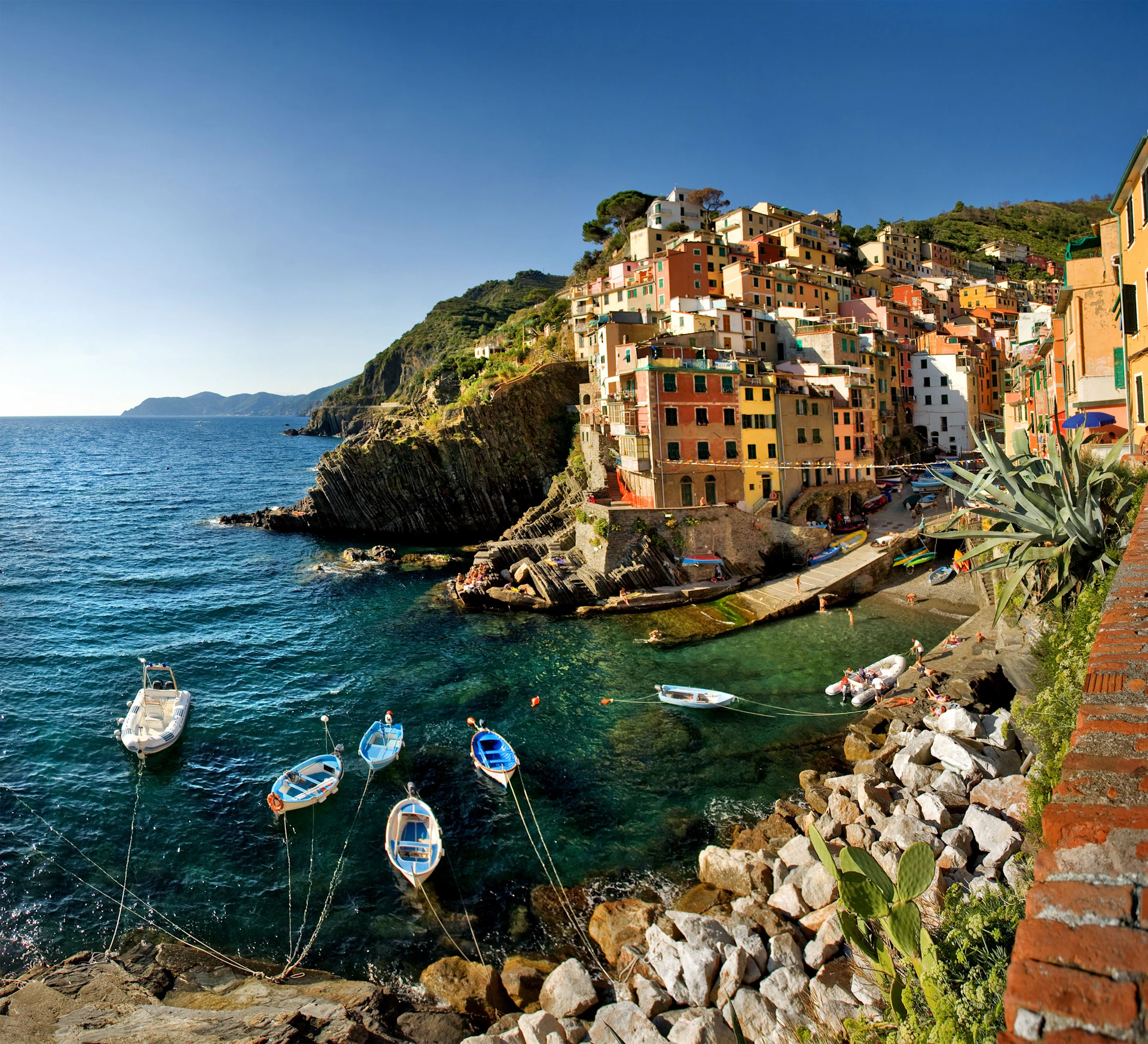 Colourful houses on a cliffside beside a calm blue sea with several small boats tied to the shore. Rocky terrain in the foreground.