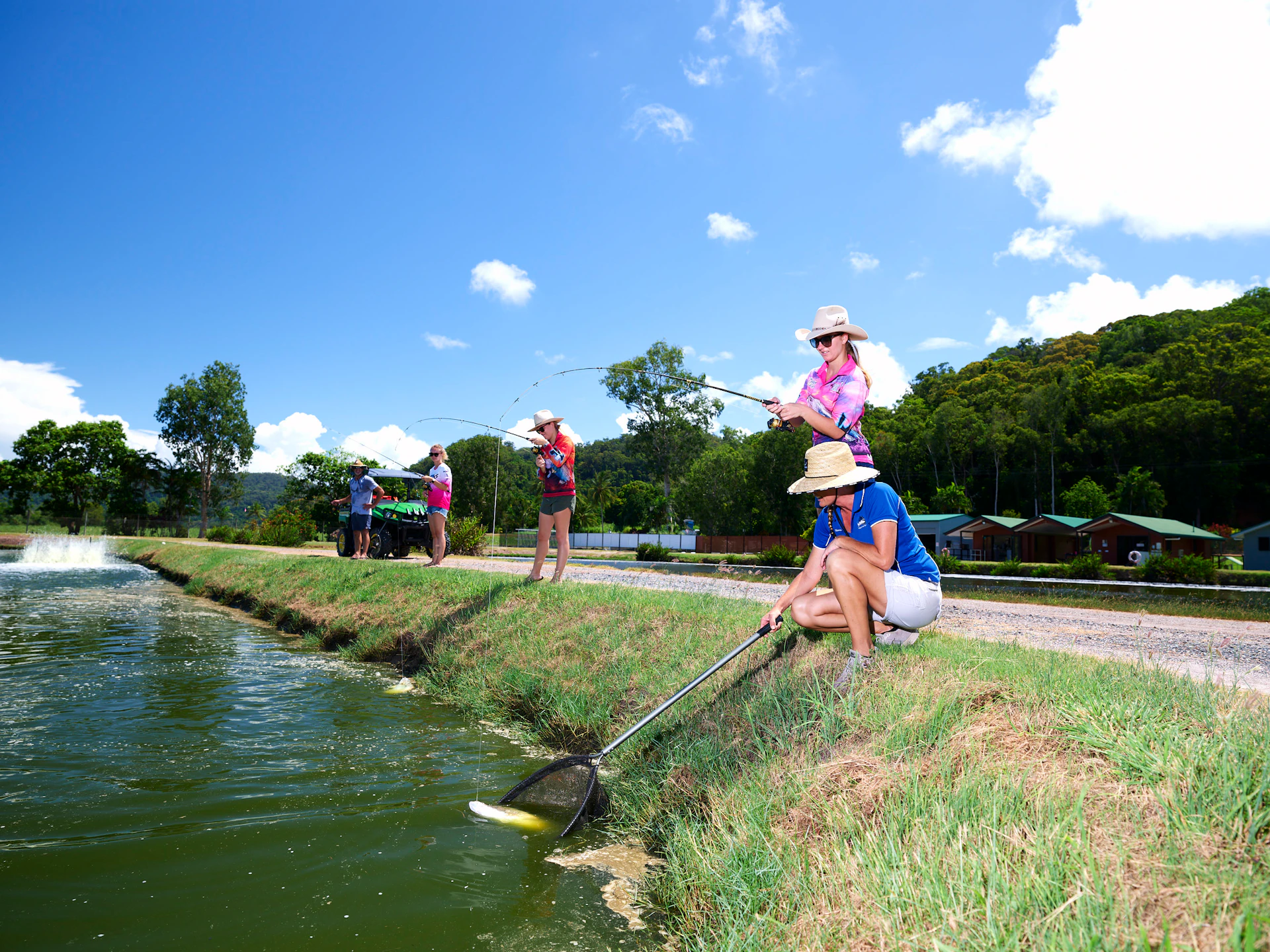 A group of people fishing by a pond under a clear blue sky. One person kneels with a net, while others stand with fishing rods.