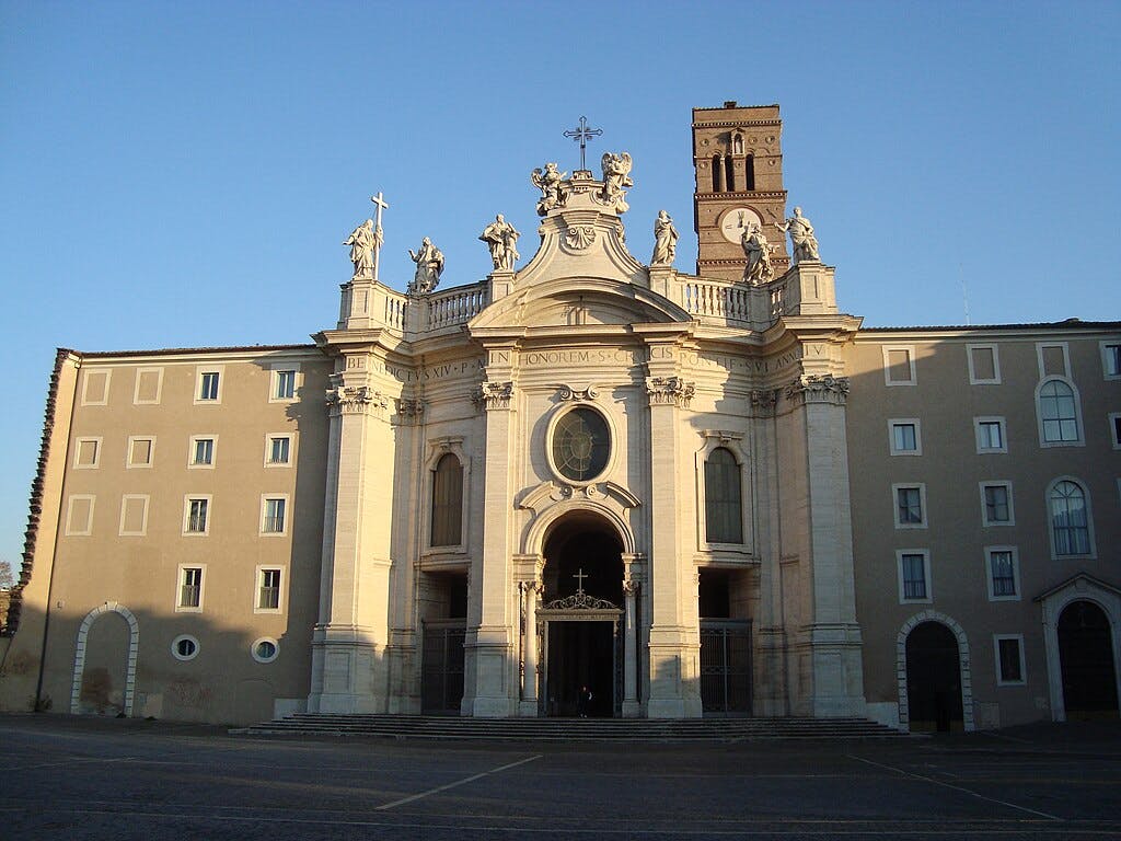 Ornate church façade with statues, arched entrance, and a clock tower, flanked by symmetrical, rectangular buildings at sunset.