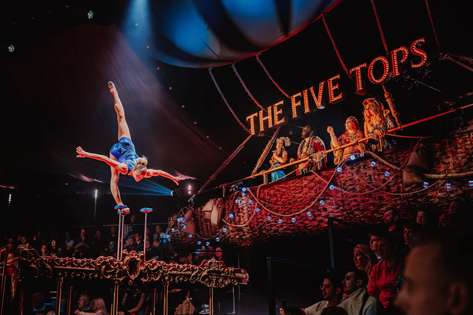 An acrobat performs on hand-balancing props in a circus tent while spectators watch from ornate seating labeled "The Five Toes."