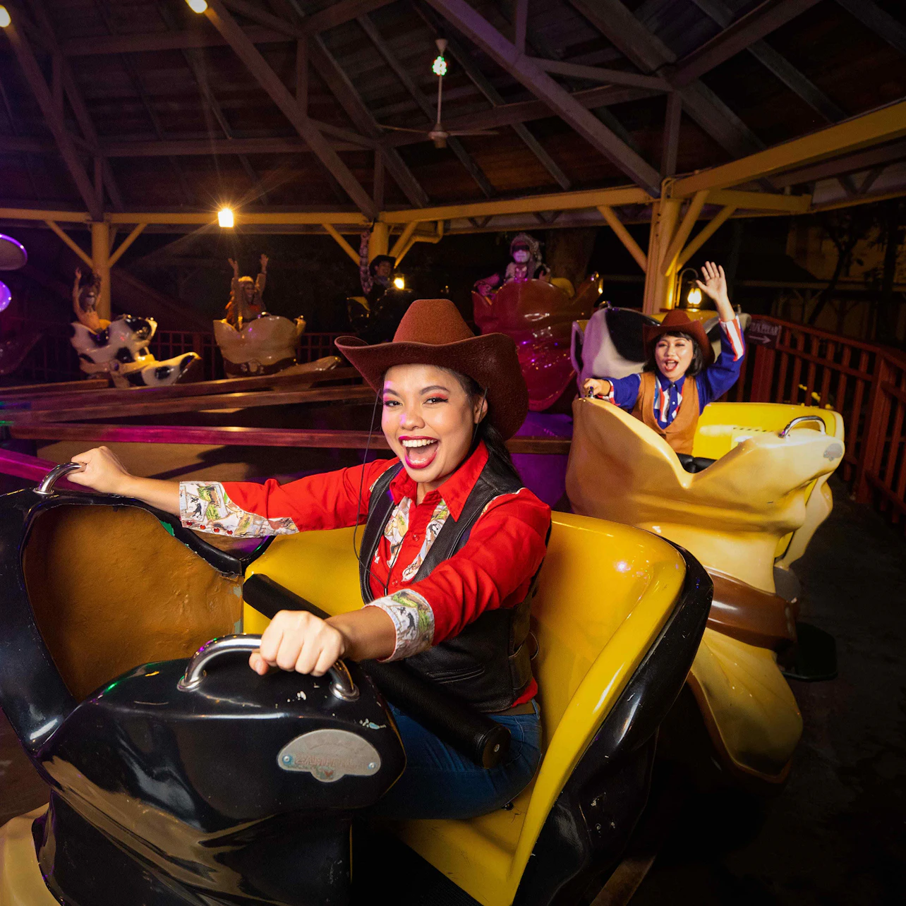 People dressed as cowboys enjoying a spinning amusement ride with bright lights in a wooden pavilion at night.