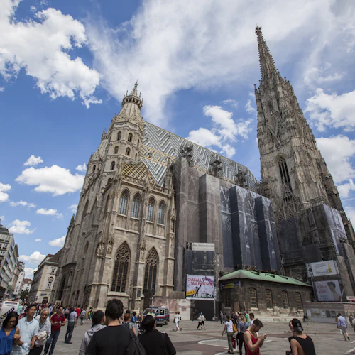 Folkmassa som går nära en katedral i gotisk stil med en hög spiral, delvis täckt för renovering, under en blå himmel med moln.